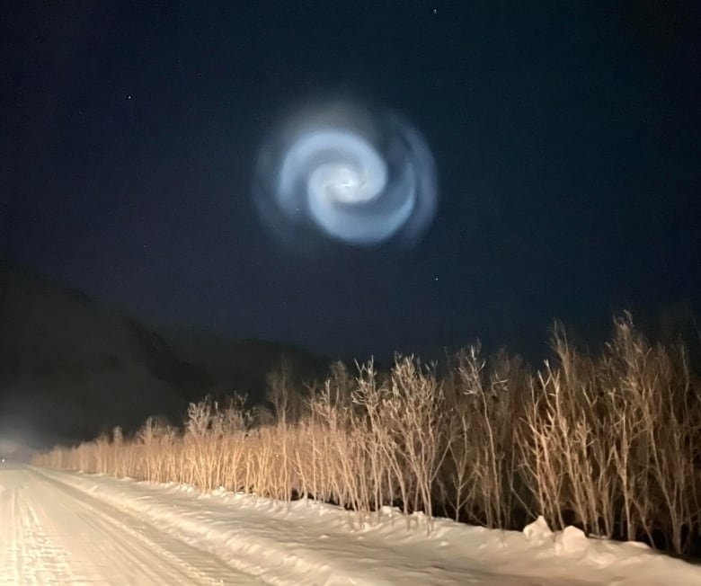 A bright swirl is seen in the night sky over a snowy road in the mountains.