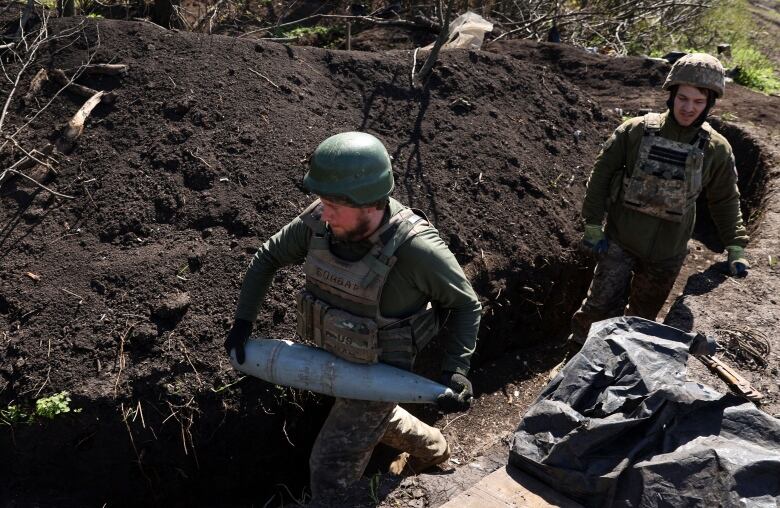 A pair of men in military fatigues are shown in a trench, with one of them carrying a shell.