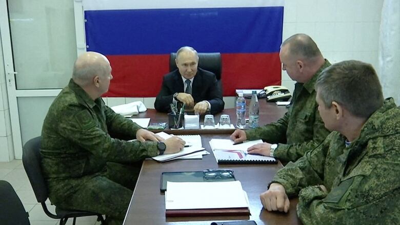 A man in a suit is shown seated at a table with three men in military fatigues.