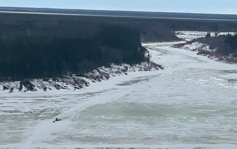 Aerial photo showing a vast wilderness, a wide mostly-frozen river and a snowmobile out on the ice. 