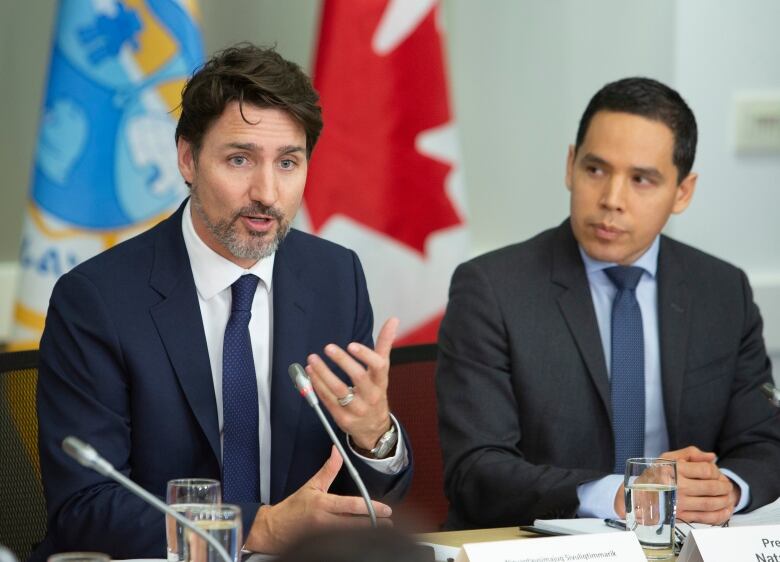 Two men in suits sit at a desk with microphones and glasses of water. The one on the left is speaking and gesturing.