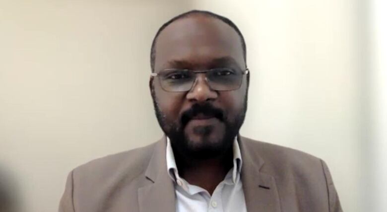 A man in a tan suit and glasses looks into the camera. 