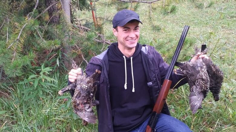 A man holds two dead birds with a riffle leaning against his knee.