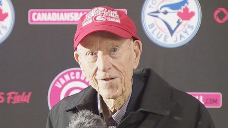 An older man in a baseball cap with Canadians baseball and Toronto Blue Jays decals behind him looks at the camera with a boyish grin