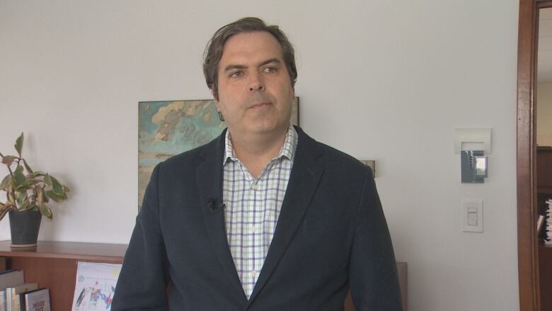 A man with short brown hair stands in front of a wall. He is wearing a blazer with a shirt.