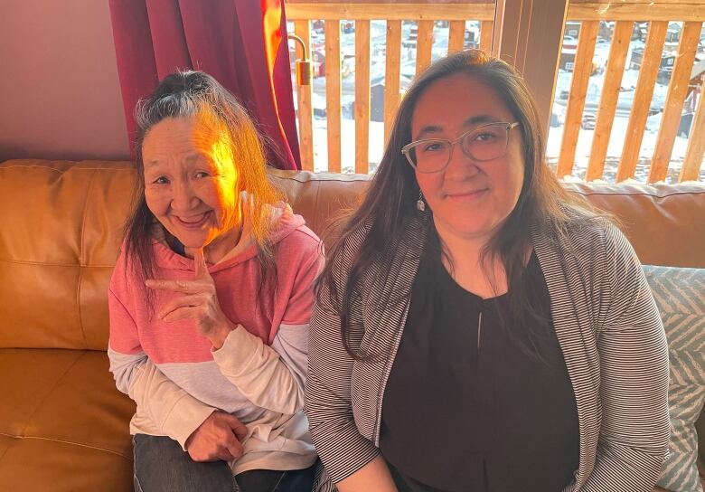 Two smiling women sit on a sofa in front of a window. 