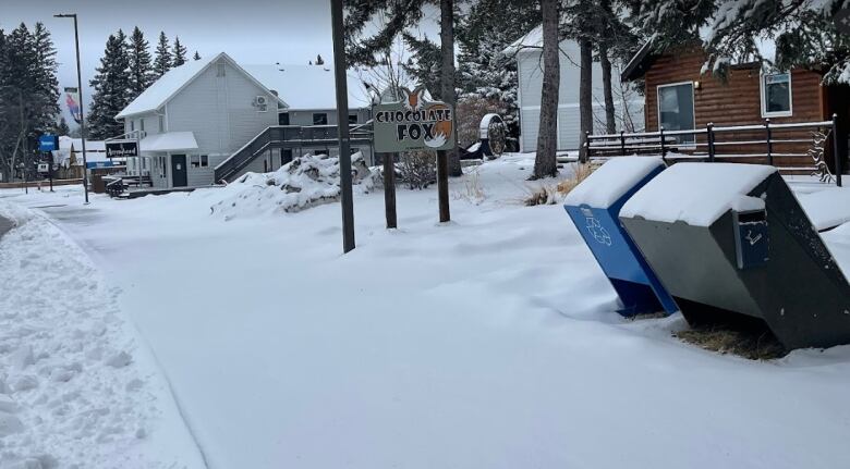 A snowy sidewalk and road in Riding Mountain National Park. Garbage bins, signs and building roofs are all covered in a fresh layer of snow.