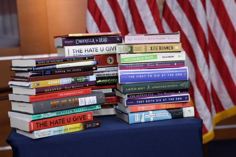 Three stacks of books on a table, covered in a blue cloth, in front of red and white striped flags in the background.