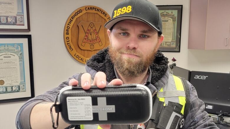 A man in a black cap and yellow high visibility vest holds up a naloxone kit.