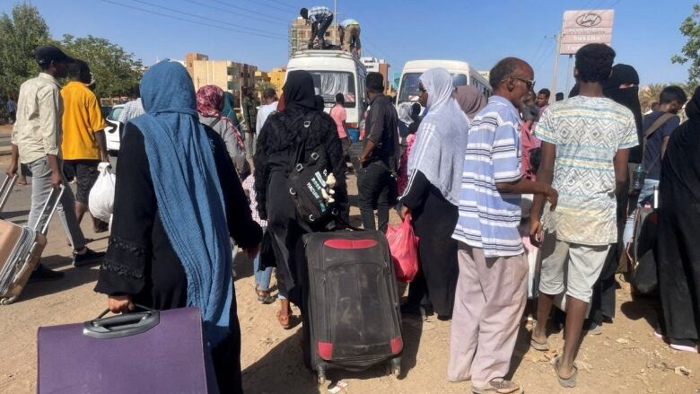 A crowd of civilians, many carrying luggage, gather on a busy road. 