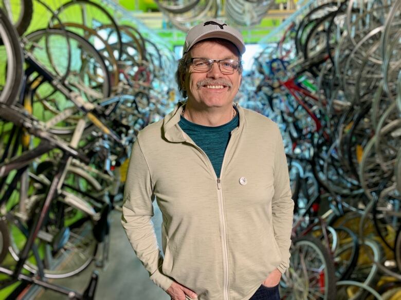 A man in a ball cap and mustache stand in-between a sea of bikes in a shop. 