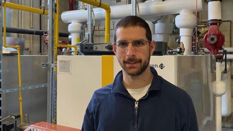 A smiling man with short dark hair, a beard and glasses standing in front of an HVAC system.