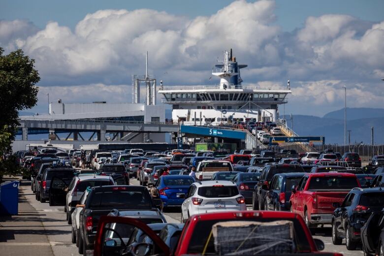 Hundreds of cars are waiting for a B.C. Ferry on a sunny day. The docked ferry is framed by the ocean and mountains.