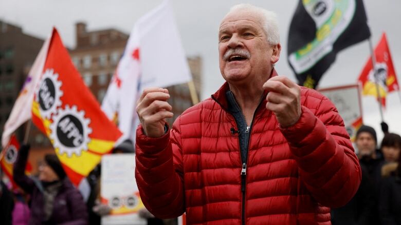 A union leader speaks to a rally. People hold various flags behind him.