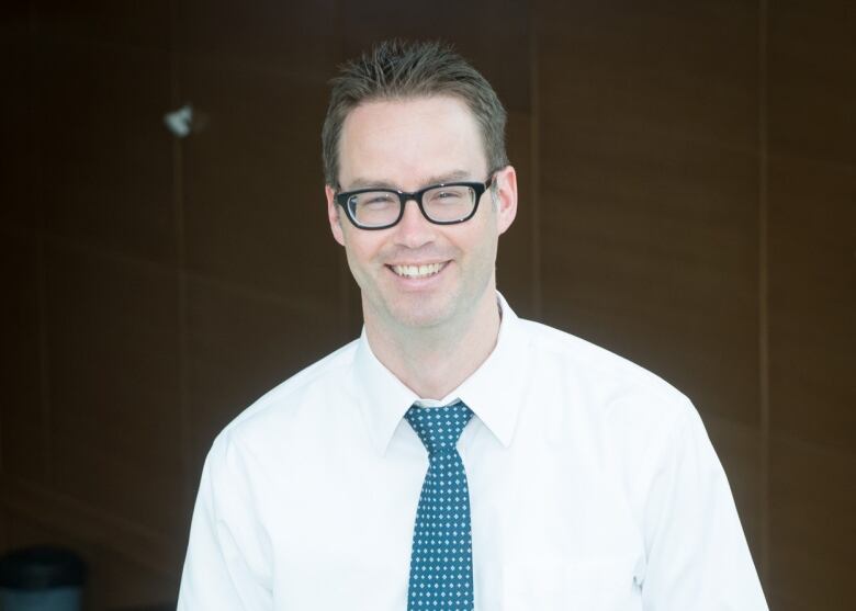 A smiling man in a white shirt with a blue tie and glasses leans against a railing.