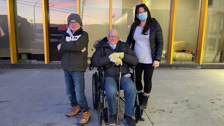 A man sits in a wheelchair smiling next to a young boy and a woman, who is wearing a mask. 