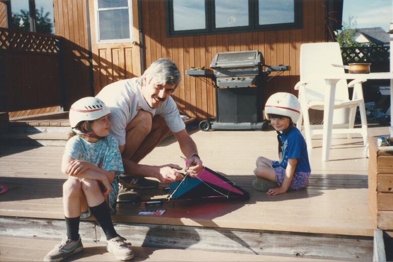 A man holds a kite between two children wearing helmets sitting on a wood deck. 