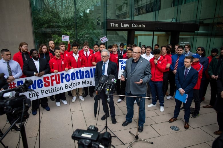 Alumni and supporters of the Simon Fraser Football program are pictured outside of the courthouse in Vancouver, on April 13, 2023. Many wear red jackets, and some hold a large sign that says Save SFU Football.