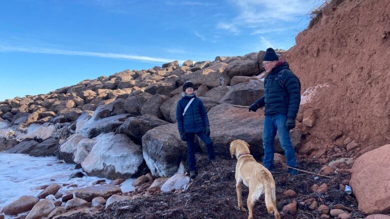 A man and woman and a dog stand near a pile of large boulders while the sea water hits against the stones.