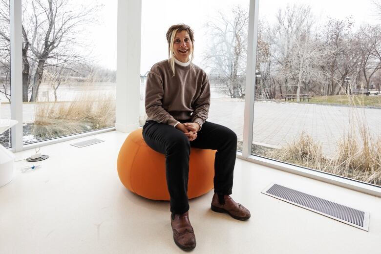 A woman sits on an orange bean bag chair in a hotel lobby surrounded by glass with a view of a river and a trail in the background.