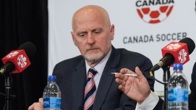 A bald man wearing a suit and tie sits at a table with a microphone in front of him.