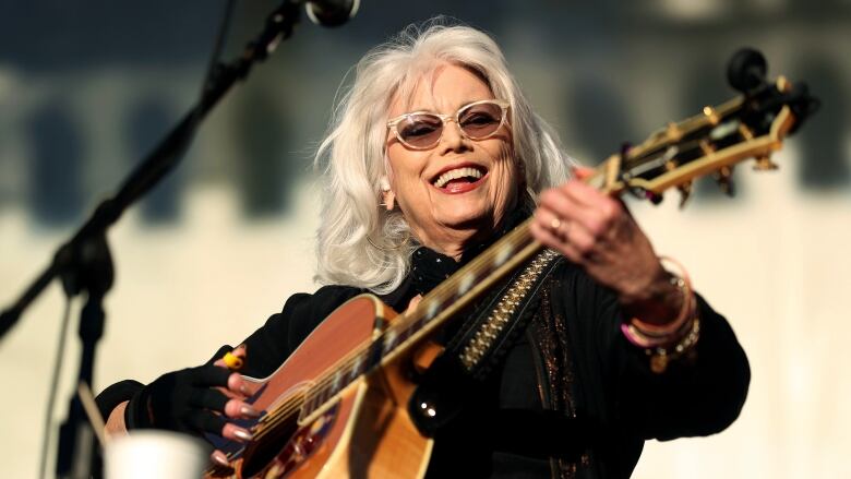 Wearing sunglasses and a dark shirt, a white-haired woman smiles while playing an acoustic guitar.