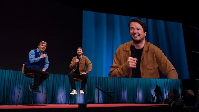 Two men speak to each other while seated on high chairs on a stage. A screen displaying the face of one of the men is above them. The man's face on the screen looks like the other man's face.