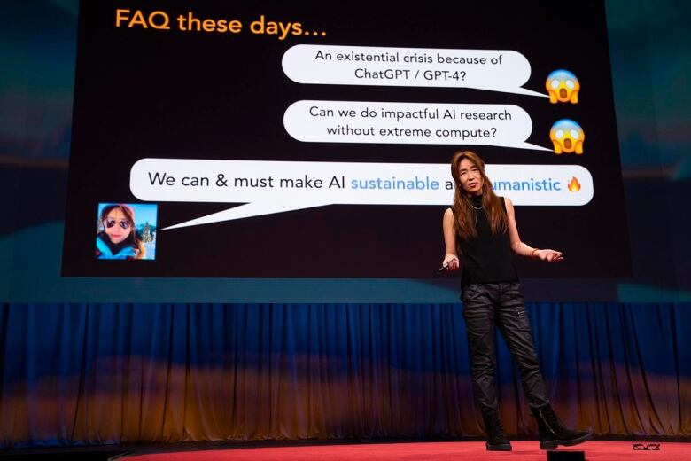 A woman dressed in black stands on a stage with a social media dialogue displayed on a screen behind her.