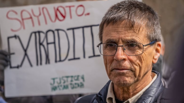 A man with glasses stands in front of a sign which says 