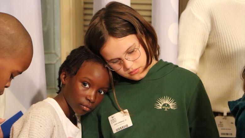 A little girl leans her head on the shoulder of another girl.