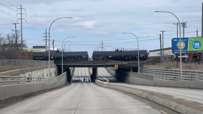 A black tanker from a train leans out from a concrete bridge above a street.