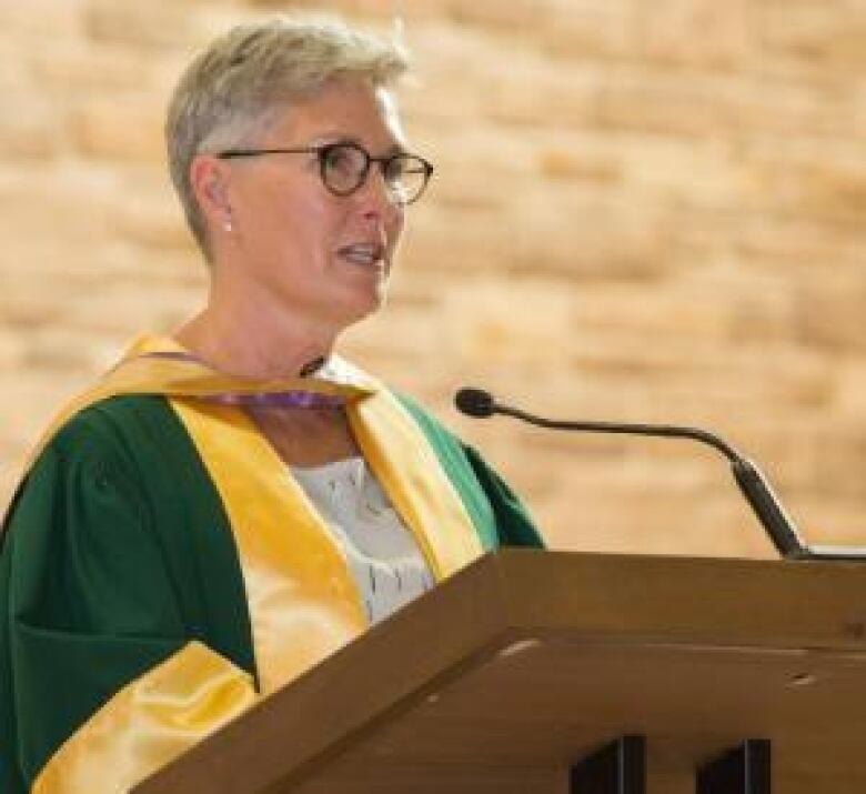 A woman with short hair and glasses wearing a green and yellow robe, speaking into a microphone at a podium.