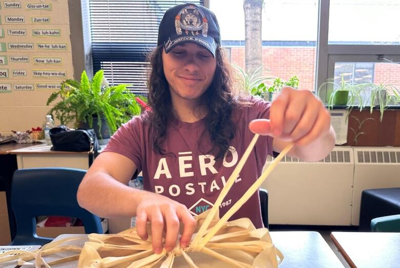 A young man wearing a red t-shirt and a baseball cap sits at a desk, looping his finger through laces attached to deer hide in the shape of a circle.