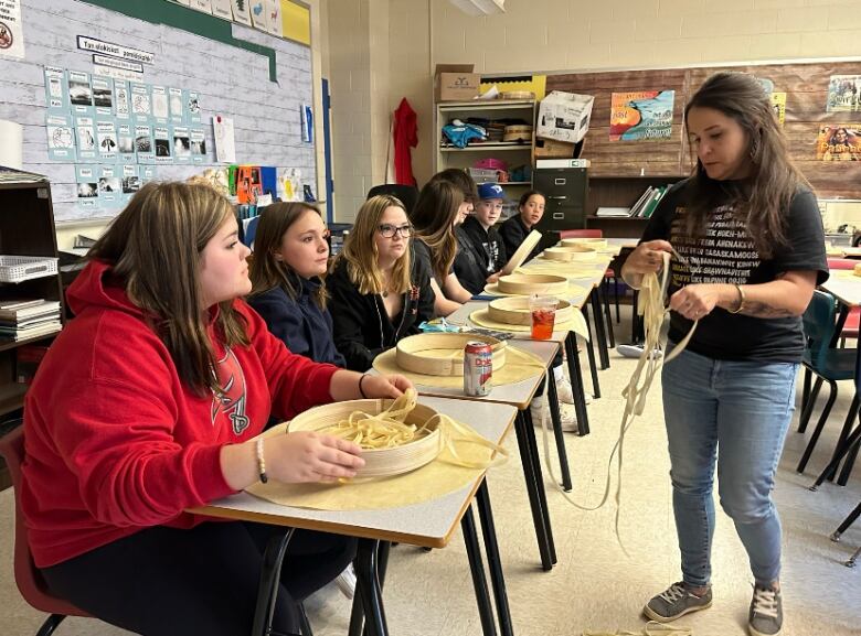 A group of students sit at a row of desks, each with lace and deer hide in front of them for drum-making. An woman stands in front of them, holding some lace.