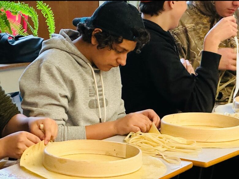 A young man sits at a desk, bent over some lace that's stringing through deer hide.