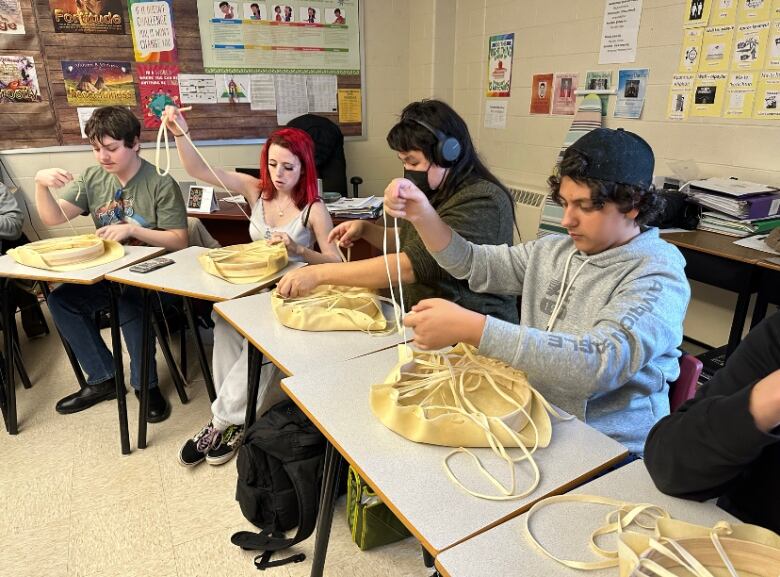 High school students sit in a row of desks, each in different stages of pulling lace through deer hide to make drums.