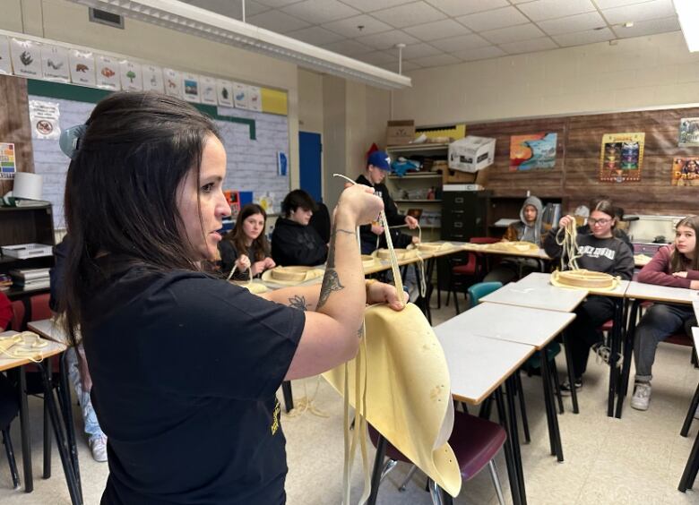 A woman stands in a classroom in front of a row of students, holding up lace and deer hide.