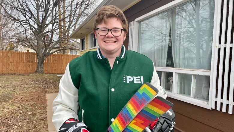 Para Hockey player Aidan Godin smiles for a picture. He is wearing a green jacket that says P.E.I. and hockey gloves. Godin is holding his para hockey sledge and sticks tapped in rainbow colours. 