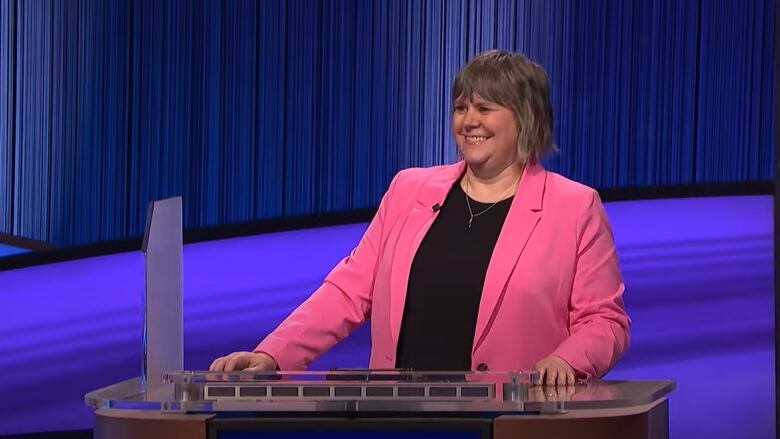 A woman wearing a pink blazer smiles broadly as she stands at a game show podium.