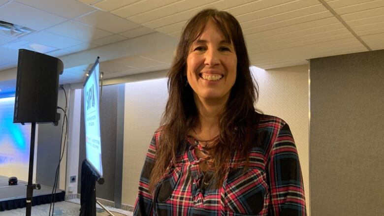 A woman with long dark hair smiles to the camera as stands in a hotel conference room.