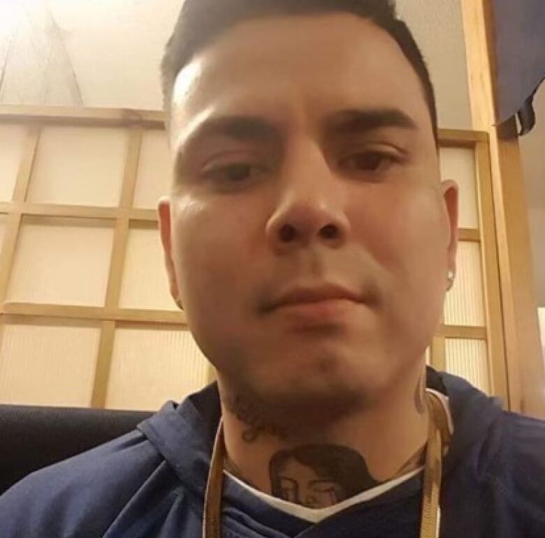 An Indigenous man with a tattoo on his throat is wearing a lanyard and a blue hoodie. He has black hair and a clean-shaven face.