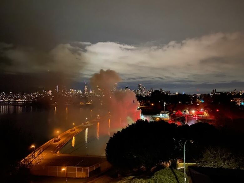 Plumes of smoke rise from near a swimming pool, with Vancouver's skyline visible in the background.