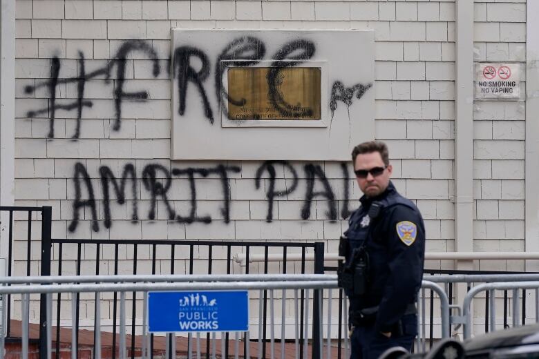 A police officer in uniform and dark glasses standings near a metal barricade adorned with a blue sign that says, 