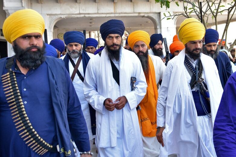 A group of bearded men in turbans walk. The man at the centre of the photo and a man in the foreground who has a bandolier slung over his shoulder eye the camera, apparently warily. 