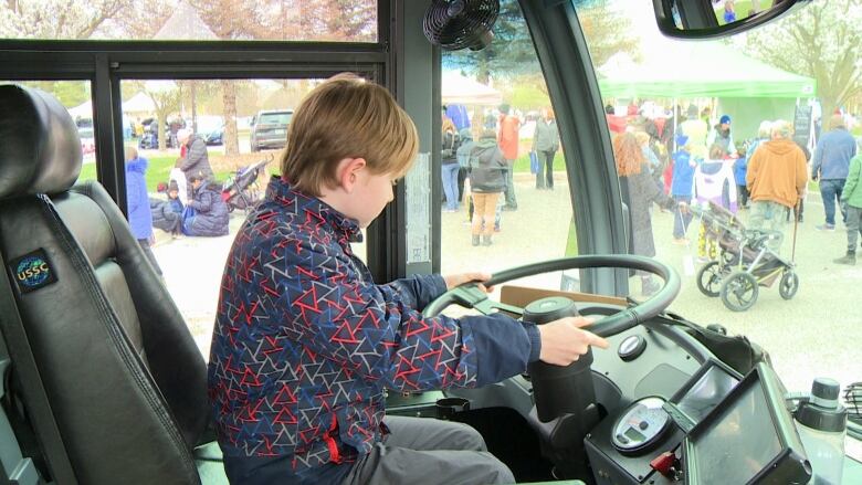 A child wearing a black jacket with blue red and grey triangles sitting in the driver's seat of a stationary bus