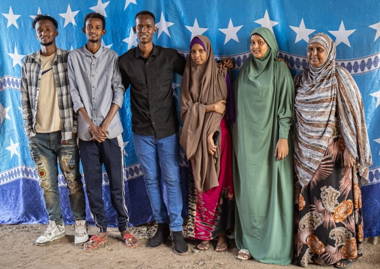 Three young men, two young women, and their mother, pose with their arms around one another in front of a blue curtain on which there are white stars.