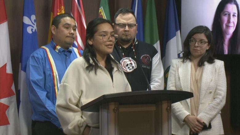 Girl with shoulder length brown hair, circular glasses and off white pullover stands at a podium. Behind her are two men and a woman. One of the men wers a ble shirt with multicoloured suspenders, the other wears a black and white bomber jacket. The woman has glasses and a white pantsuit. 