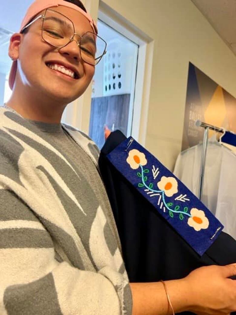 An Indigenous person holds up a blue costume, which has Wolastoqey motifs and flowers.