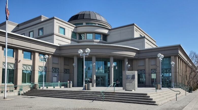 A sprawling concrete building topped by a round dome in the middle and a curved glass front with a concrete roof supported by pillars with wide steps leading up to the doors accented by double-globed lighting on poles. 