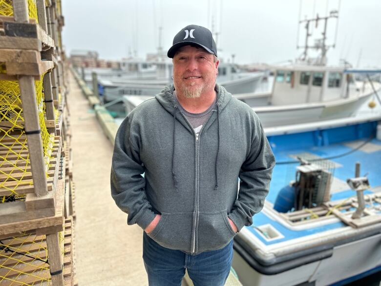 A man in a grey sweatshirt and ballcap stands next to lobster traps on a wharf 